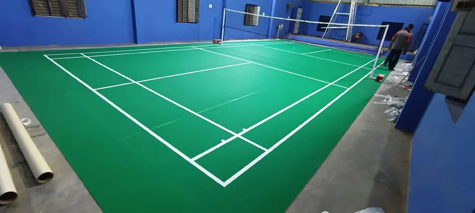Green badminton court mat freshly laid in a blue-walled hall, with installation materials beside it.
