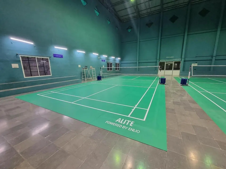 Green badminton court mat laid over a stone floor in a teal-walled indoor hall, with a second court alongside.