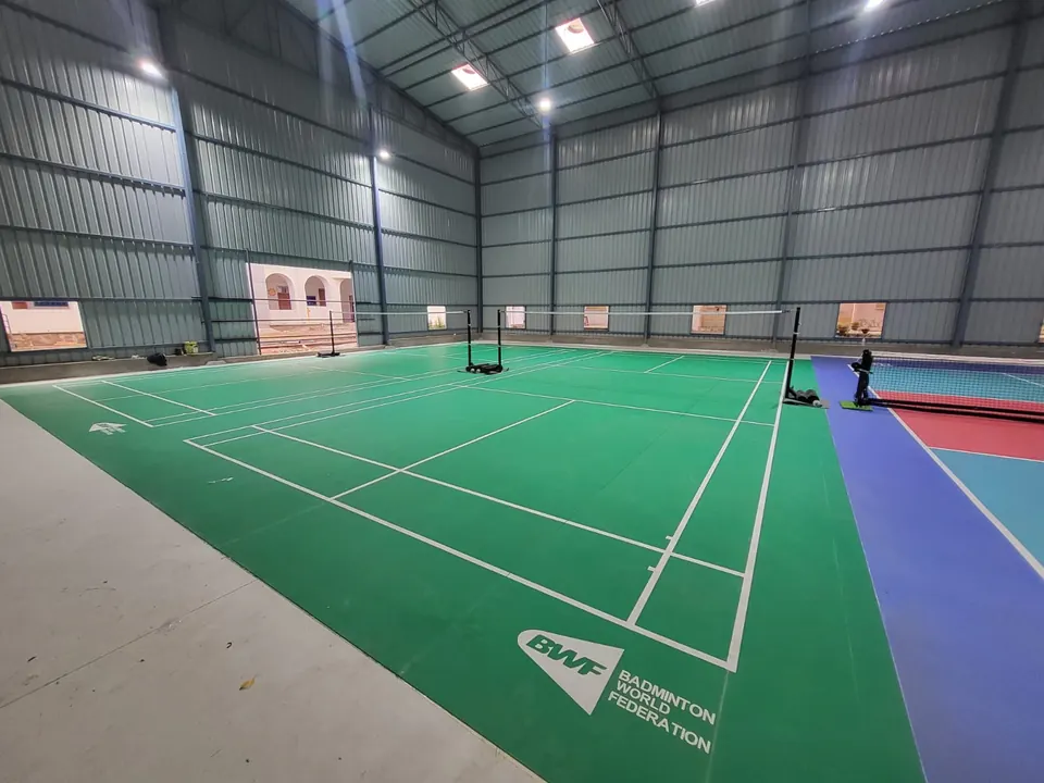 Green badminton court mats next to a blue and red pickleball court in the same steel shed.