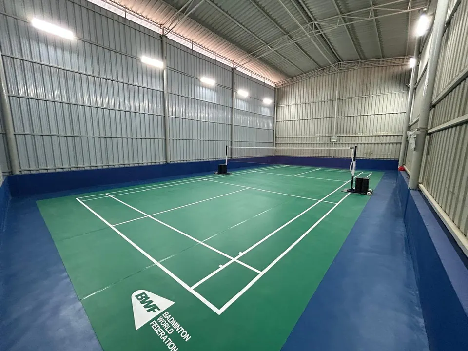 Indoor badminton court with a green court mat, white lines and a blue surround inside a steel-clad hall.