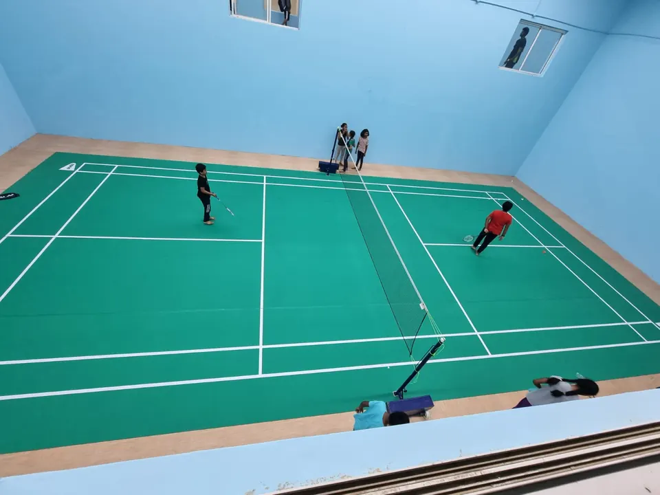 Overhead view of children playing on an indoor badminton court with a green mat and wood-finish surround.