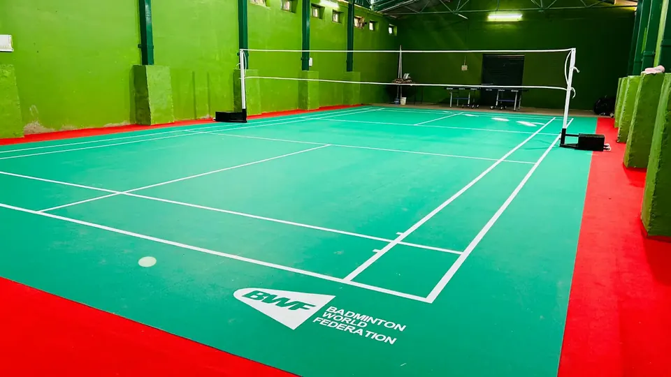 Indoor badminton court with a green mat and a red surround in a hall with green walls.