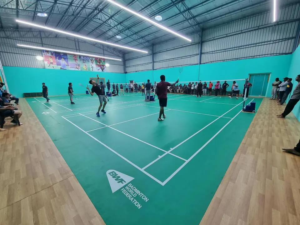 Players and spectators on the indoor badminton courts in Kalwakurthy, Telangana, built by Kelysports: green court mats with a wooden surround in a teal-walled steel hall.