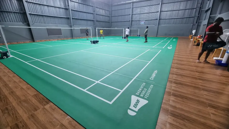 Players on green badminton court mats bordered by wood-finish flooring in a grey steel hall.
