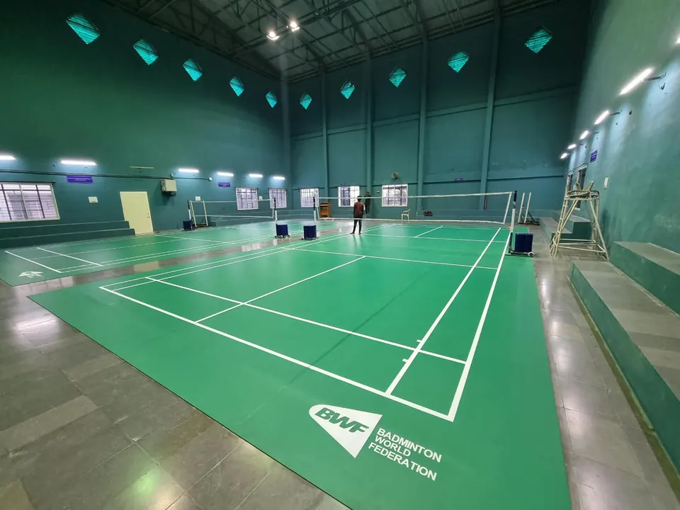 Two indoor badminton courts on green mats in a tall teal hall with an umpire chair and spectator steps.
