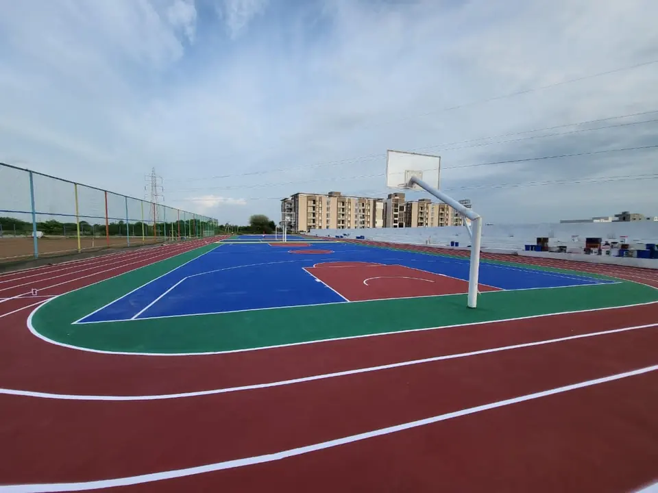 Ground-level view of the lanes of a red acrylic synthetic running track curving around an outdoor acrylic basketball court with a pole and backboard.