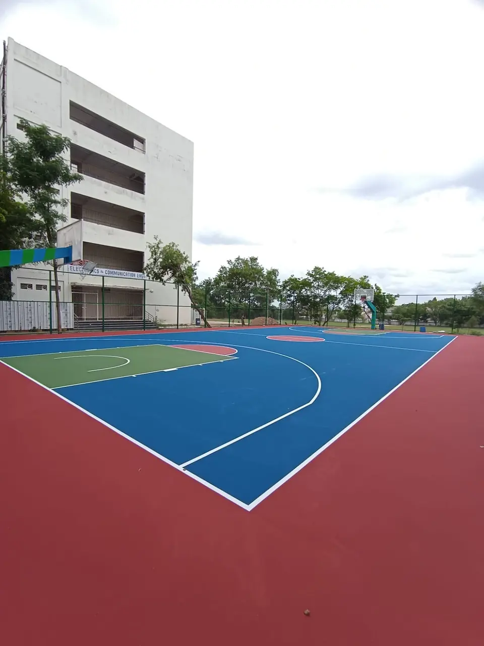 Outdoor acrylic basketball court in blue with a red surround and green key, next to a multi-storey building.
