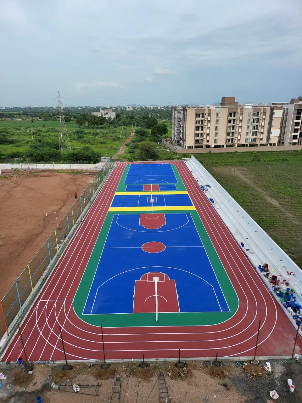 Aerial view of an outdoor acrylic basketball court with yellow multi-sport lines, surrounded by a running track, beside apartment blocks.