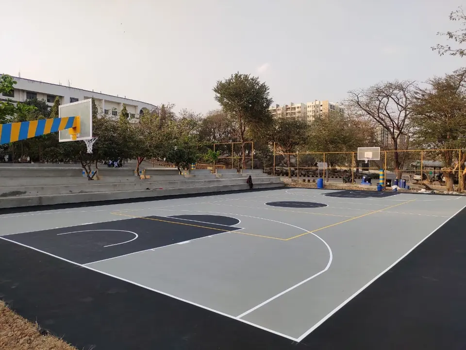 Outdoor acrylic basketball court in charcoal and grey with white and yellow lines, poles at both ends and spectator steps alongside.