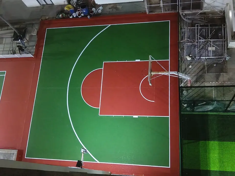 Overhead view of a half basketball court in green with a red key and surround, with the pole and backboard fitted, next to an artificial turf area.