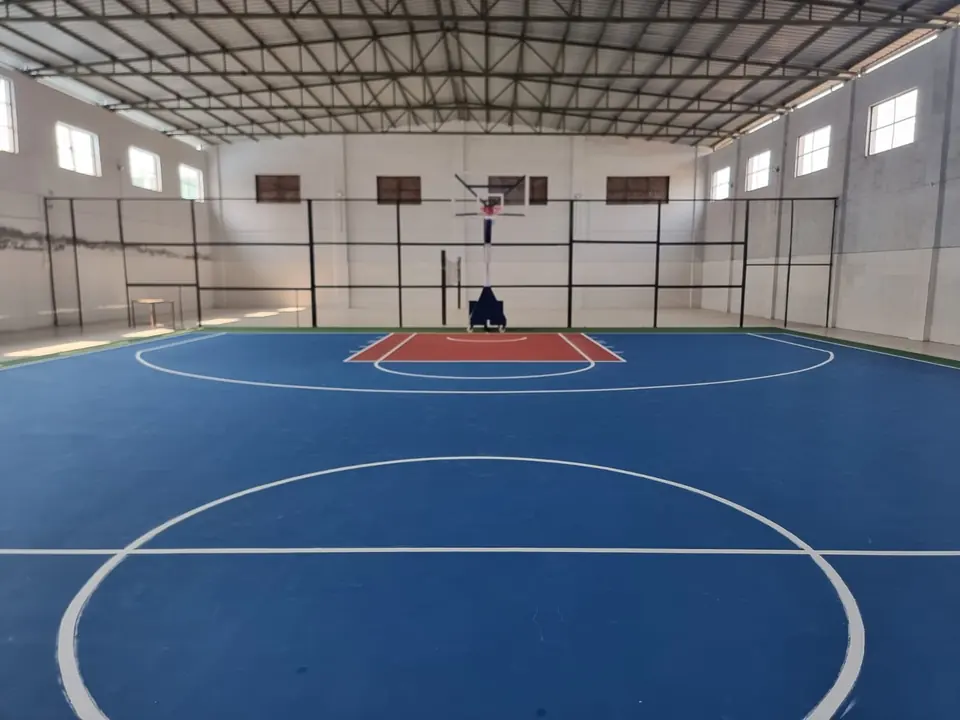 Indoor basketball court with a blue playing surface and red key inside a hall with a steel roof, high windows and a pole at the far end.