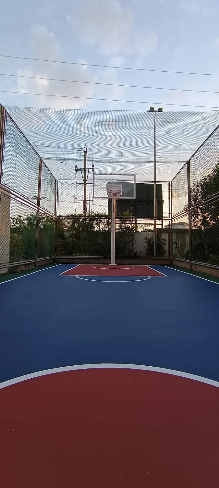 Outdoor acrylic basketball court in blue and red enclosed by tall netting, with a pole and backboard and a floodlight pole behind.