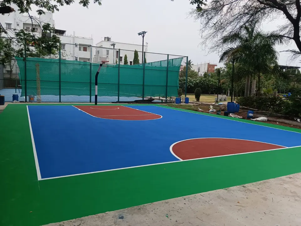 Outdoor acrylic basketball court in blue with red keys and a green surround, with green shade netting along one side and poles at both ends.