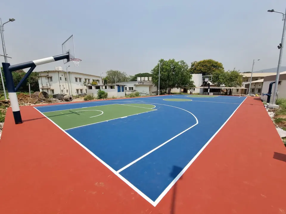 Outdoor acrylic basketball court with a blue playing area, green key and red surround, with a black and white basketball pole in the corner.