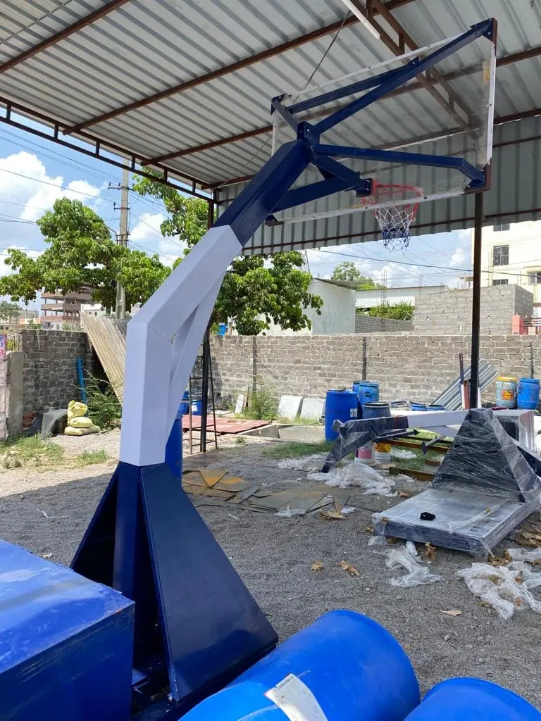 Angled blue and white boom of a Kelysports basketball hoop system holding a framed transparent backboard, ring and net, under the workshop shed.