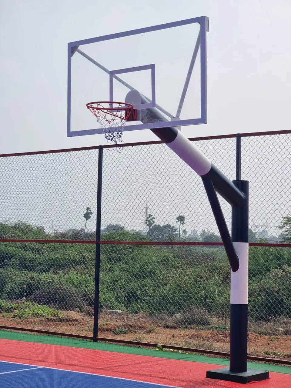Black and white basketball hoop system by Kelysports with an angled arm, braced transparent backboard, ring and net, beside an interlocking tile court.