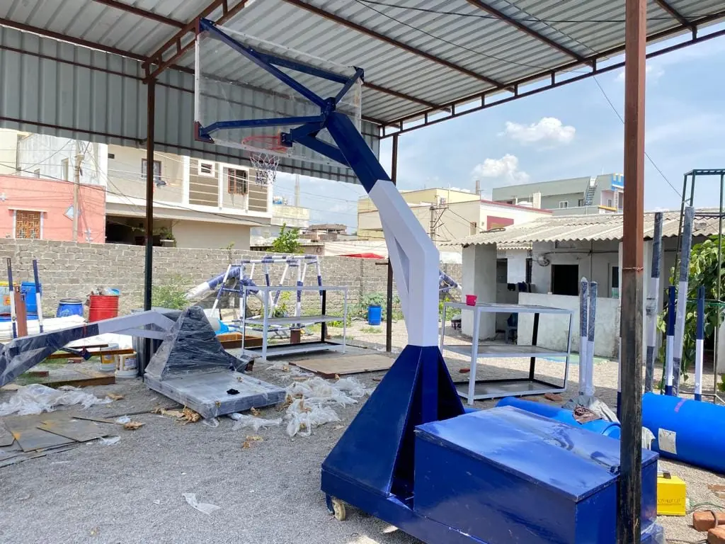 Blue and white basketball hoop system with a wheeled base box, angled boom and braced backboard, finished in the Kelysports workshop yard.