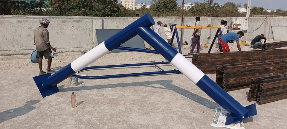 Freshly painted blue and white Kelysports steel pole frames with base plates, laid out during fabrication while the team works on steel sections nearby.