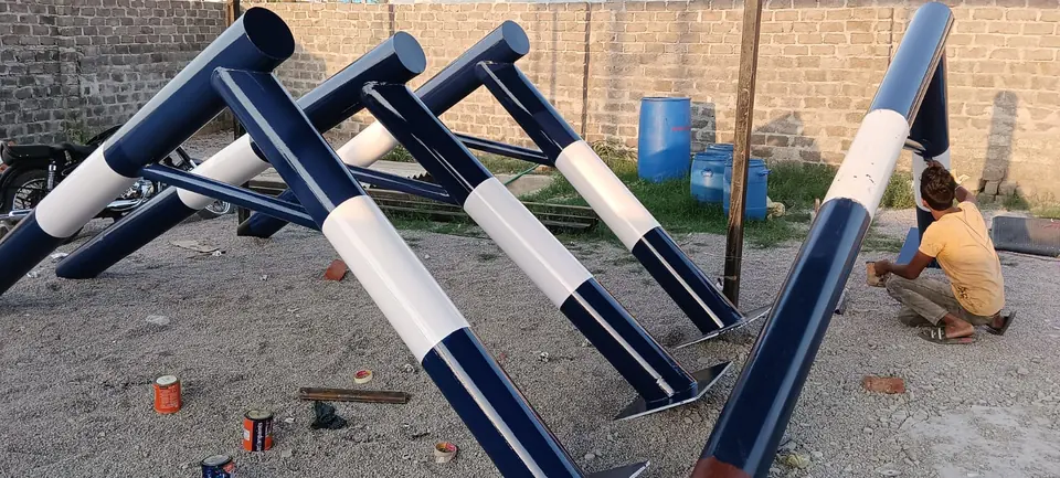 Several Kelysports basketball pole frames in navy blue and white standing in the workshop yard while a worker finishes the paint by hand, with base plates welded on.