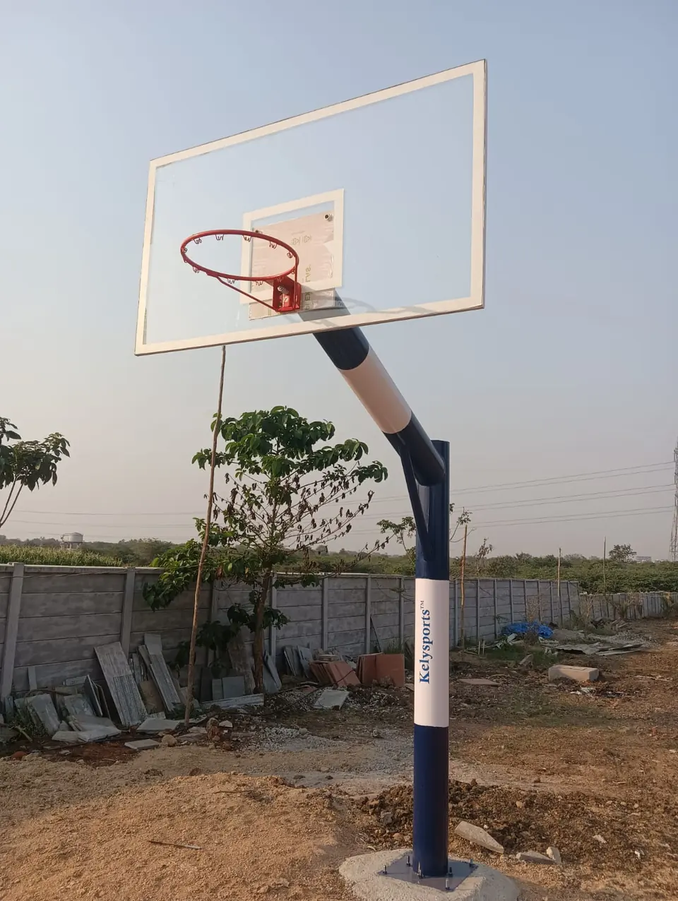 Newly installed Kelysports basketball pole with its base plate bolted to a concrete footing, the backboard still in its protective film.