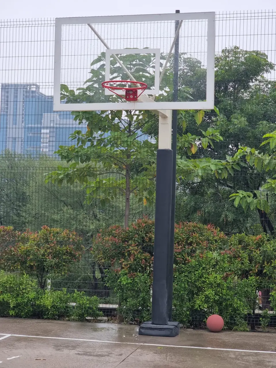 Kelysports basketball pole with a padded post and padded base, a framed transparent backboard and red ring, beside a mesh-fenced outdoor court.