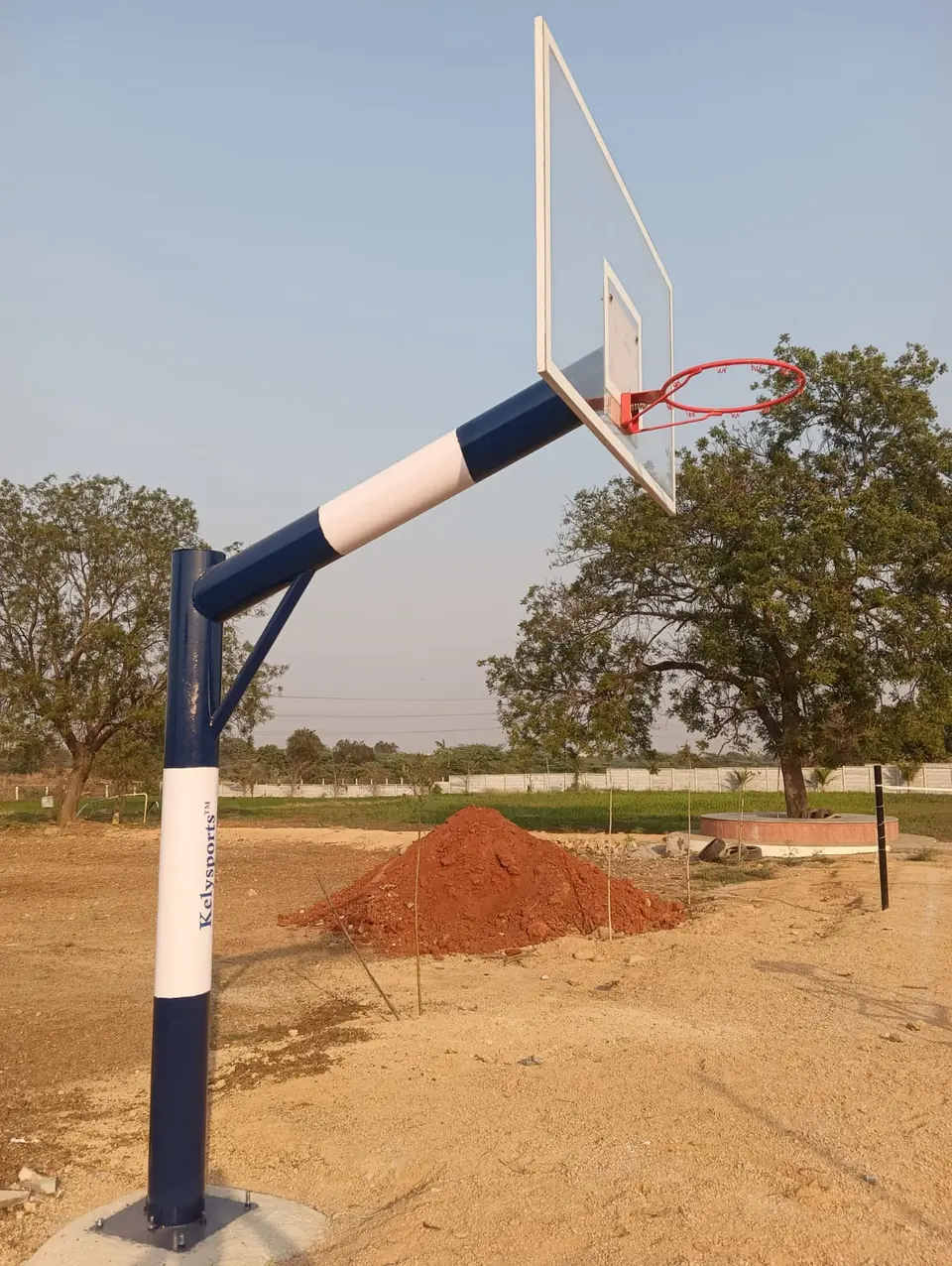 Blue and white Kelysports-branded basketball pole, made in the Kelysports workshop, with an angled arm, transparent backboard and red ring, bolted to a concrete footing.