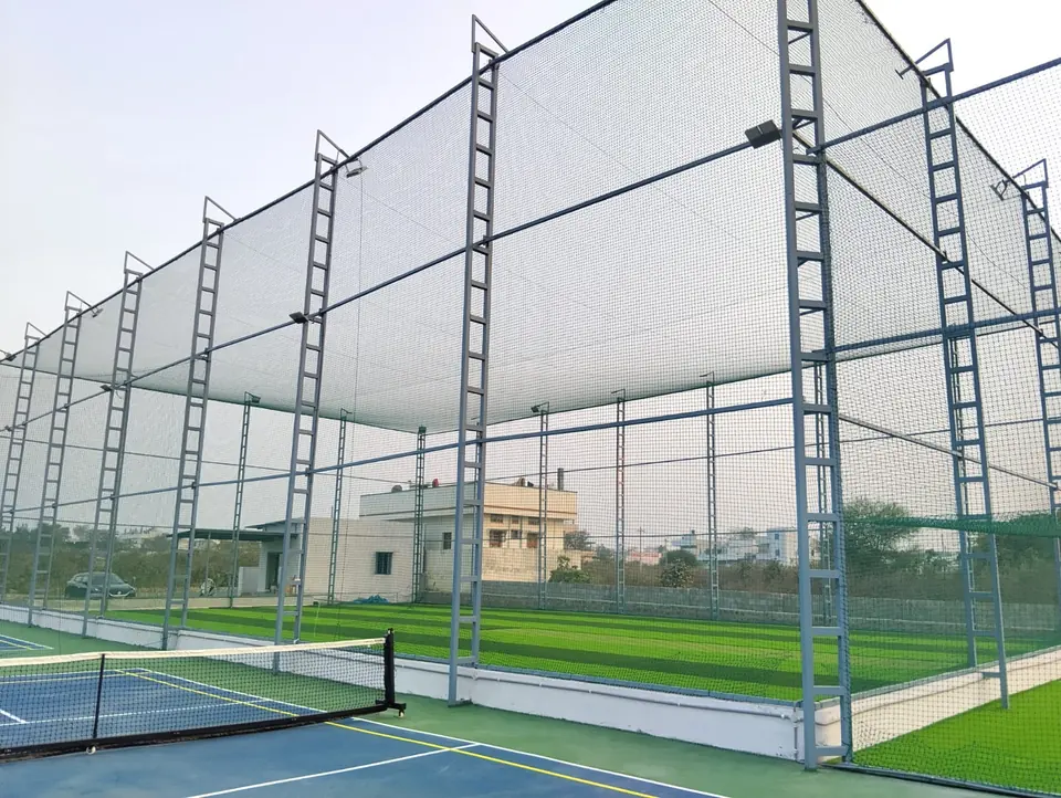 Tall box cricket net enclosure on grey ladder-frame steel poles with floodlights and artificial turf, next to a pickleball court with its net.
