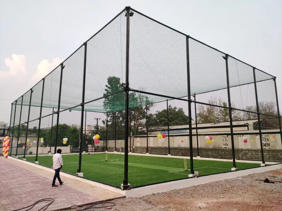 Box cricket court construction by Kelysports: a large netted arena on black steel poles with floodlights, artificial turf, a cricket pitch, stumps and white field markings.