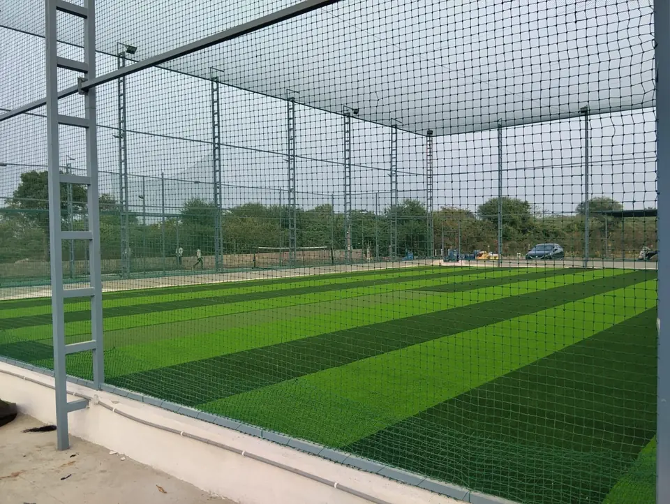 Box cricket court with striped artificial turf inside a tall net enclosure on grey ladder-frame steel poles, with roof netting and floodlights.