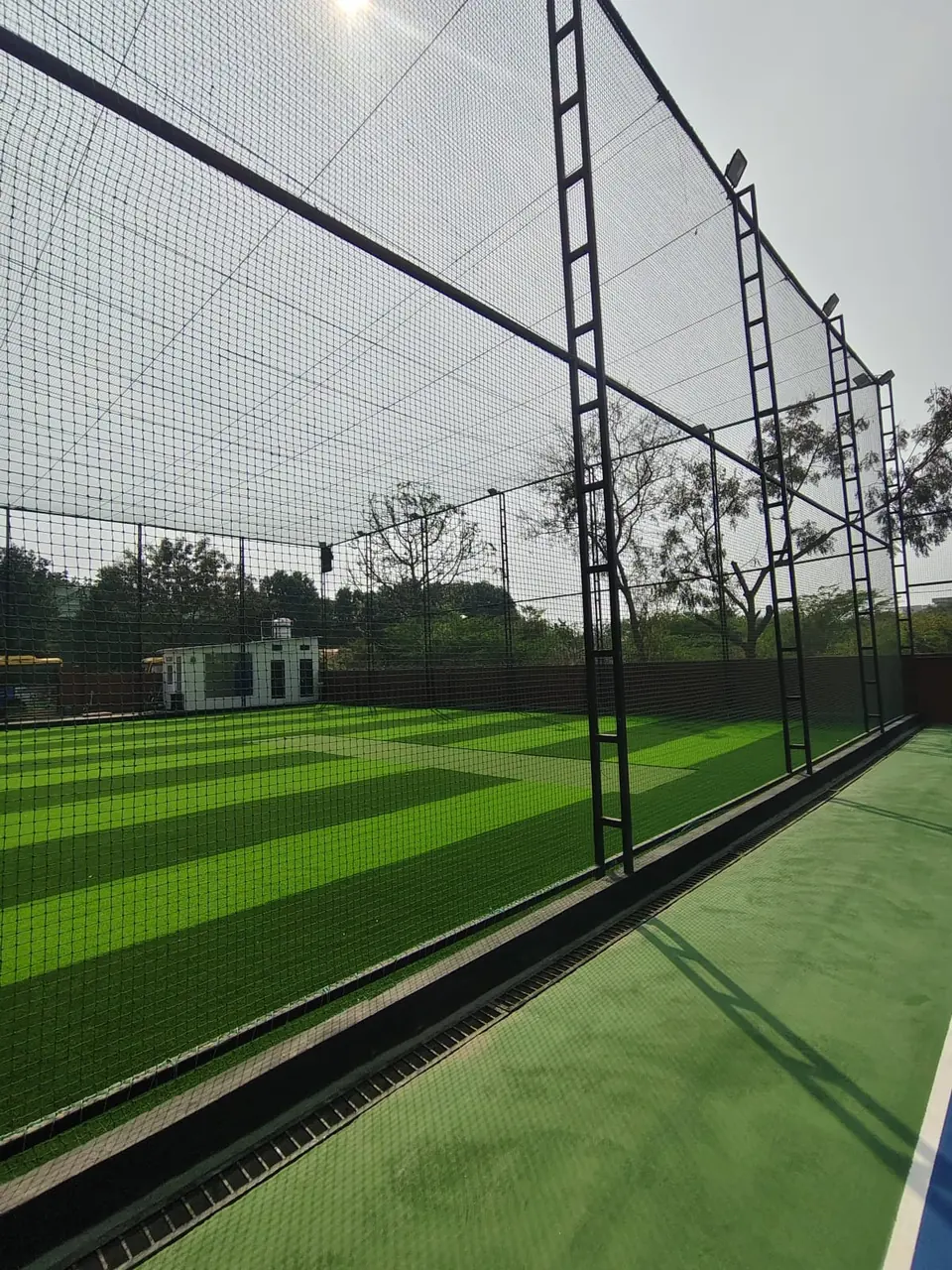 Box cricket arena with striped artificial turf and a lighter cricket pitch strip, enclosed by netting on black ladder-frame poles with floodlights, beside a hard court.