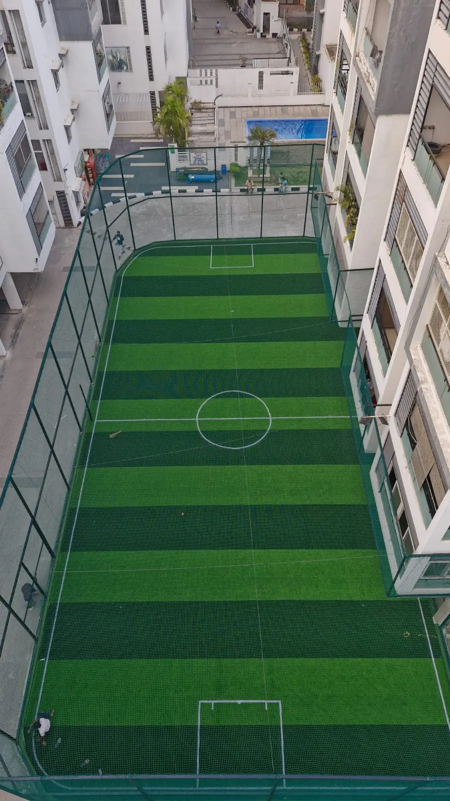 Aerial view of a netted football turf ground with goal areas, a centre circle and striped artificial grass, set between residential towers beside a pool.