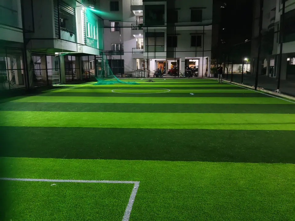 Football turf with white line markings and a centre circle, lit at night inside a netted enclosure between apartment blocks.