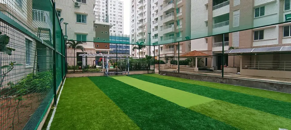 Netted artificial turf court in a residential community with a cricket pitch strip and a small football goal, surrounded by apartment towers.