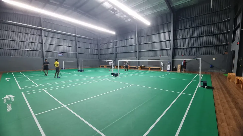 Completed indoor sports hall by Kelysports: green badminton court mats with posts and nets, players on court, linear LED lights and wood-finish flooring inside a grey steel-clad shed.