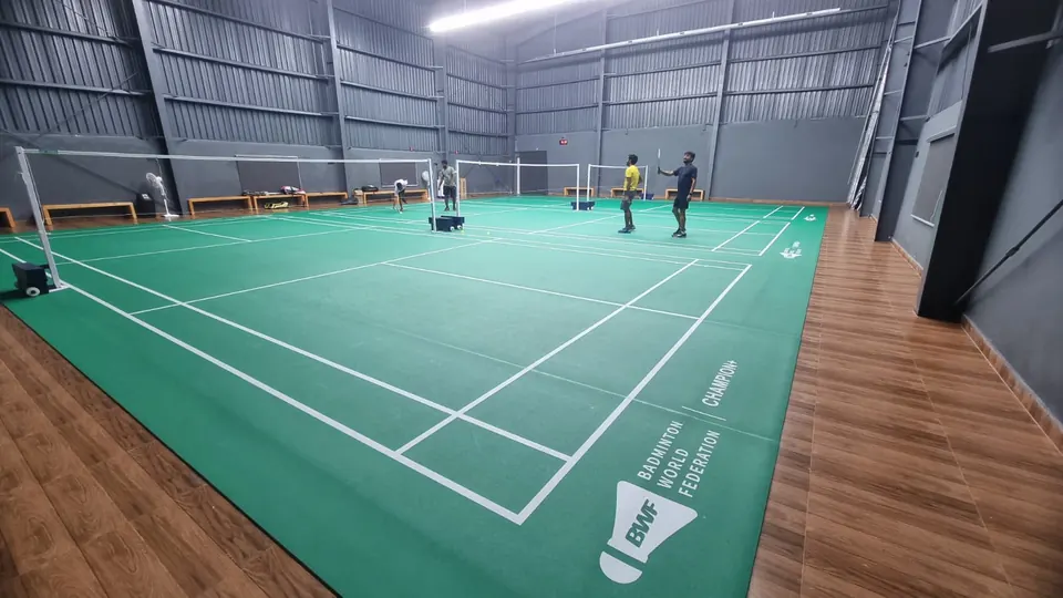 Indoor badminton courts with green court mats, white court lines, posts and nets on a wood-finish floor in a grey steel-clad hall, with players warming up.