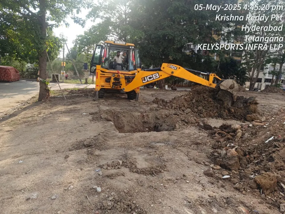 Excavator digging and clearing the ground for a new indoor sports facility, with trees and apartment buildings around the plot.