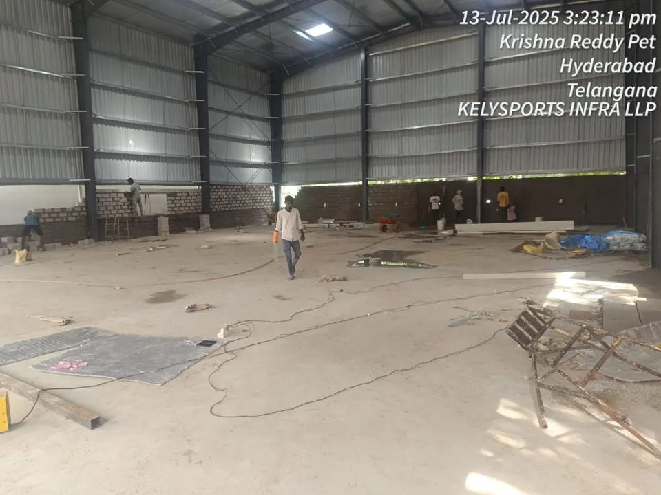 Masons building blockwork walls along the base of the clad steel shed, inside the large open hall.