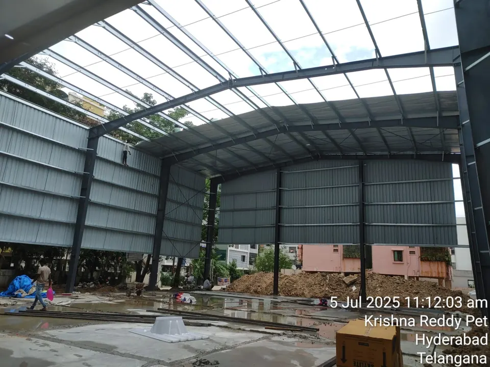 Steel portal frame of a sports shed with wall cladding fixed and roof sheeting in progress, workers on the frame.