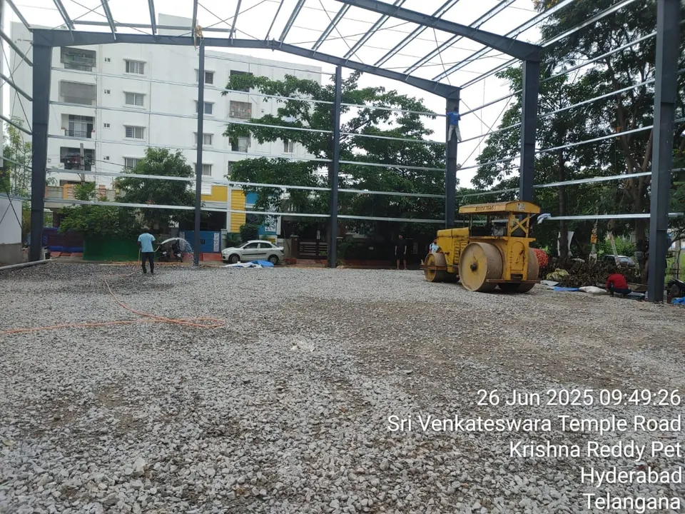 Road roller compacting a crushed-stone sub-base while a worker waters it, under the newly erected steel frame of a sports shed.