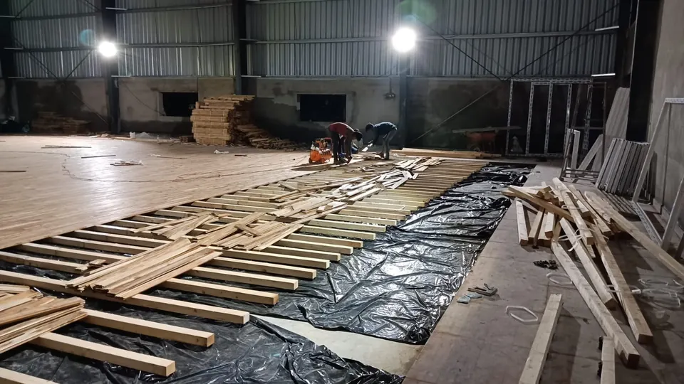 Wooden floor boards being nailed onto the battens, with the finished section of the floor behind and stacks of timber in the hall.
