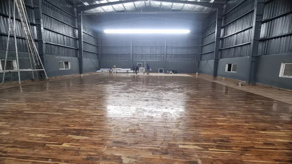 Finished, lacquered wooden sports floor reflecting the lights in a tall steel-clad hall, before court lines and equipment.
