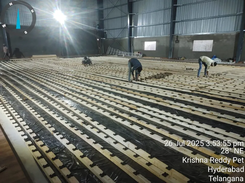 Workers fixing timber battens on cushioning pads over a black moisture barrier, the sub-floor for a wooden sports floor.