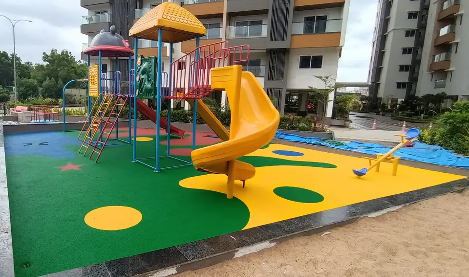 Green and yellow EPDM playground flooring installed by Kelysports, with circle and star designs beneath a playset beside apartment towers.