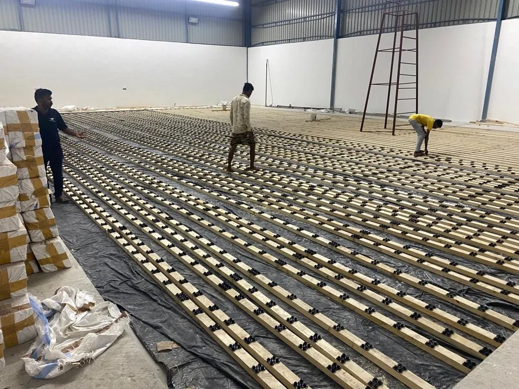 Kelysports team laying the runners and cushion pads for the Kalwakurthy badminton hall: rows of timber runners on black cushion pads over a polythene moisture barrier, with boxed flooring stock stacked alongside.