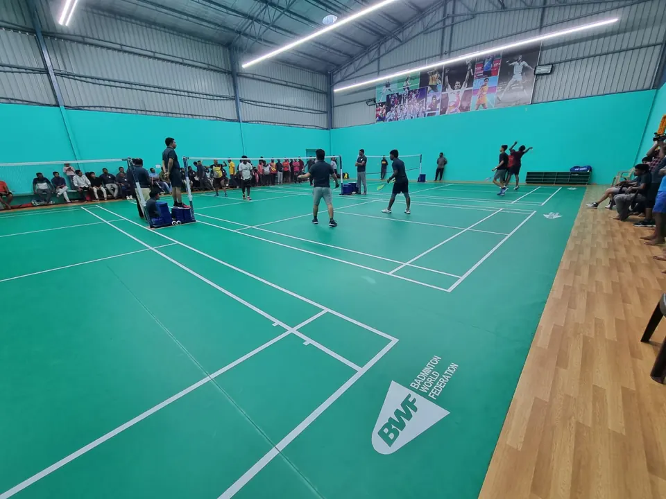 Players and spectators at the finished indoor badminton courts in Kalwakurthy, Telangana: green court mats with white lines laid over the teak wooden floor, in a teal-walled steel hall with linear LED lights.