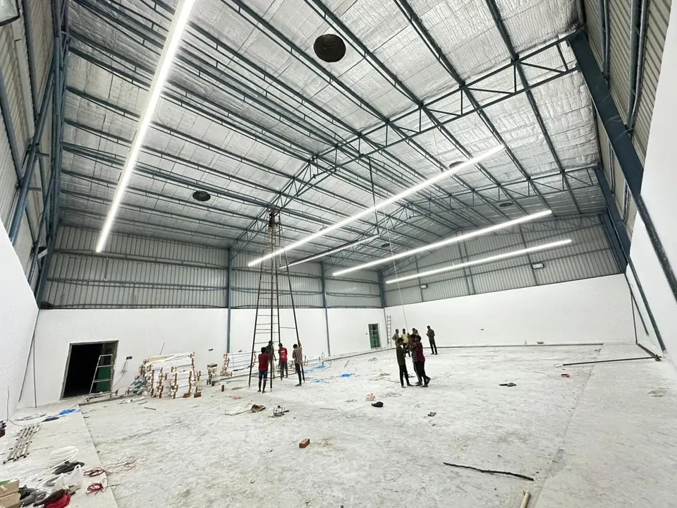 Inside the Kalwakurthy indoor badminton hall before flooring: white walls, an insulated steel roof and linear LED lights being fixed from a mobile tower above the bare concrete floor.