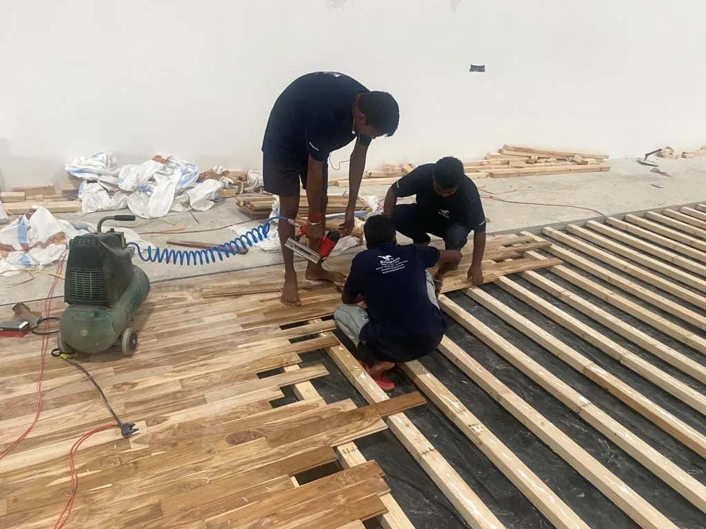 Kelysports installers nailing teak floor boards onto the timber runners with a pneumatic nailer and compressor, with the bare runners still visible beside the finished rows.
