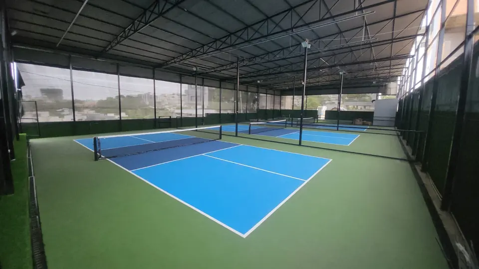 Three pickleball courts in light and dark blue with a green surround under a steel-truss roof, divided by netting and screened with mesh along the sides.