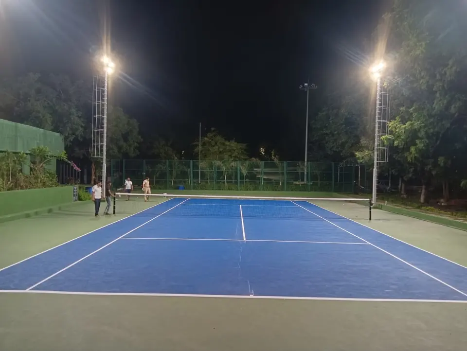 Outdoor tennis court in blue with a green surround, lit at night by floodlights on tall lattice masts, with players at the net.