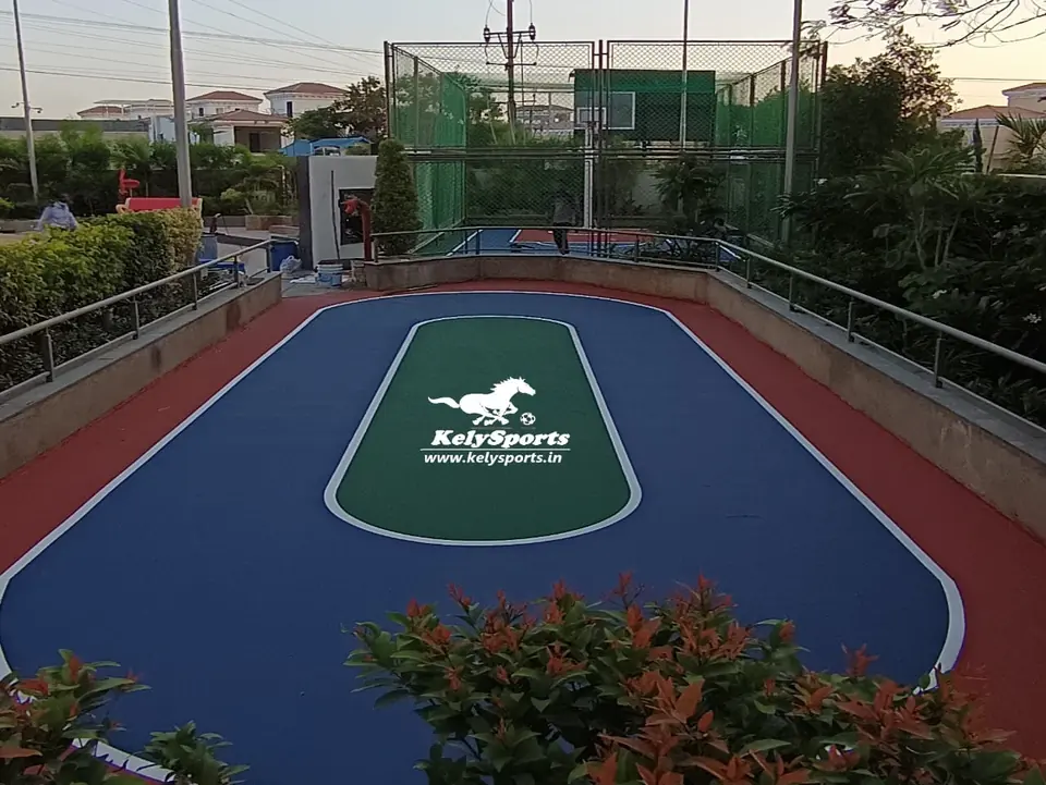 Oval outdoor skating rink with a blue track, a green centre panel carrying the Kelysports logo and a red surround, set in landscaped grounds with a netted court behind.